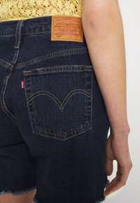 Mörkblå denimshorts med fransad nederkant, prydda med en läderlapp på bakre midjebandet och gula sömmar. Två bakfickor synliga.
