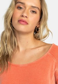 Oranssi teksturoitu yläosa, jossa leveä pääntie ja lyhyet hihat, hienovaraisella raidallisella kuviolla ja kontrastivärisillä saumauskohdilla.