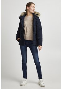 Navy blue padded jacket with a removable fur-lined hood, beige knitted sweater, dark blue jeans, and white sneakers. Simple design, casual style.