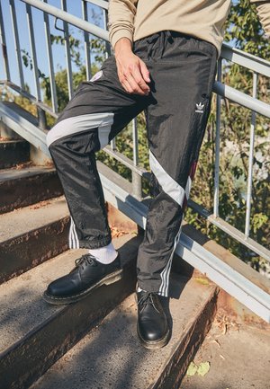 Osoba ubrana w czarne spodnie dresowe Adidas z białymi paskami i czarne buty, stojąca na metalowych schodach na zewnątrz z poręczą i tłem z liści.