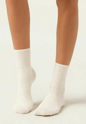 Jambes portant des chaussettes de cheville blanc cassé avec une texture côtelée subtile, debout sur une surface neutre claire avec un talon légèrement relevé.