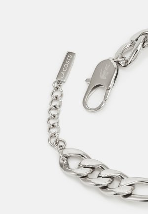 Strieborný náramok z reťaze s veľkými, prepojenými článkami. Obsahuje sponu a prívesok s gravírovaným nápisom "LACOSTE." Hladký kovový povrch.