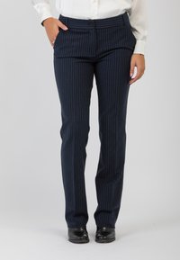 Persona che indossa pantaloni a righe blu navy, una camicetta bianca infilata dentro, scarpe nere, in piedi con una mano in tasca.