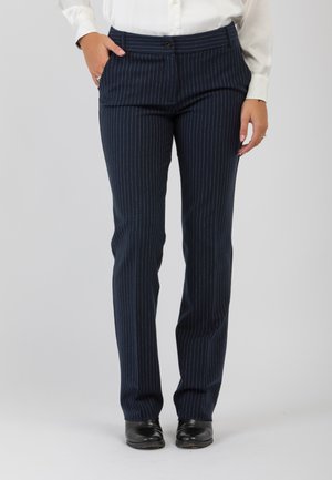 Persona che indossa pantaloni a righe blu navy, una camicetta bianca infilata dentro, scarpe nere, in piedi con una mano in tasca.