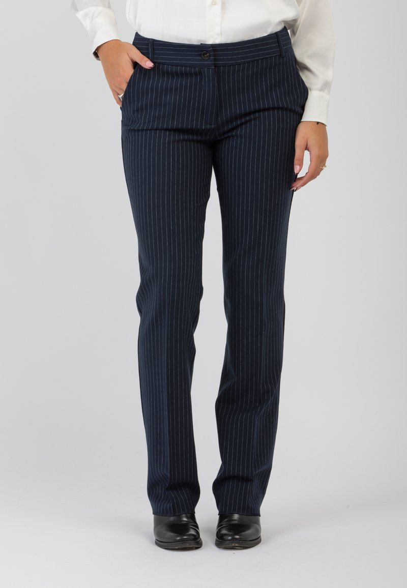 Persona che indossa pantaloni a righe blu navy, una camicetta bianca infilata dentro, scarpe nere, in piedi con una mano in tasca.