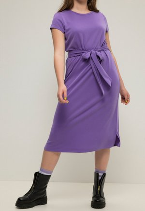 Woman wearing purple midi dress with tied waist, black zip-up boots, and matching purple socks, standing against plain background.