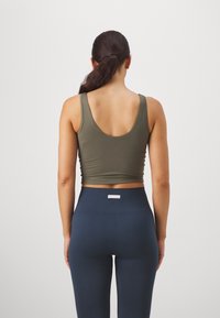 Top corto sin mangas de color verde oliva con espalda en forma de llave, combinado con leggings azul marino de cintura alta con textura acanalada y una pequeña etiqueta blanca en la cinturilla.