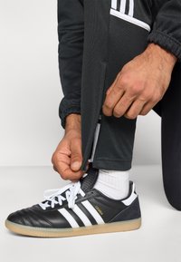 Ασπρόμαυρα παπούτσια ποδοσφαίρου με τρεις ρίγες, σόλα από καουτσούκ και υφή στο πάνω μέρος; γκρίζο αθλητικό παντελόνι με φερμουάρ στα τελειώματα και ελαστικές μανσέτες.