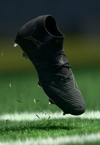 Black football boot with textured surface, featuring green accents, lace closure, and molded studs, positioned above grass with flying debris.