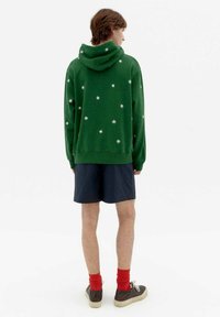 Green hoodie with white floral patterns, adjustable hood, and ribbed cuffs. Paired with navy shorts, red socks, and brown sneakers.