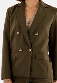 Blazer de color verde oliva con solapa doble y botones de gran tamaño, con solapas muestradas y bolsillos laterales. Tejido suave con un corte estructurado.