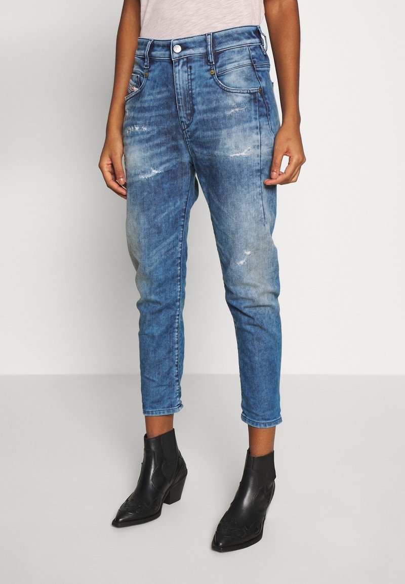 Personne portant un jean bleu délavé raccourci avec une légère usure et des bottines noires à petits talons sur fond blanc.