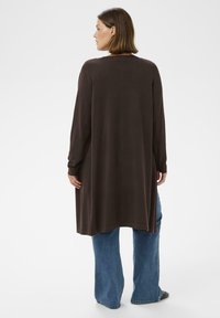 Langer brauner Cardigan aus weichem Stoff, mit einer lässigen Passform, langen Ärmeln und abgerundetem Saum, kombiniert mit hellblauen Schlagjeans.