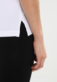 T-shirt en coton blanc avec une fente latérale présenté à côté d'un tissu noir, mettant en valeur le contraste de texture et l'ajustement sur le corps.