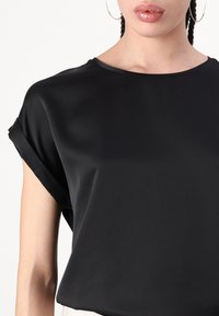 Musta lyhythihainen pusero, jossa on sileä, kiiltävä pinta ja pyöreä kaula-aukko. Käärityt hihansuut ja minimaliset muotoilutiedot.