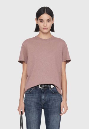 Young woman with straight dark hair wears a mauve short-sleeve shirt tucked into blue jeans with a black belt.