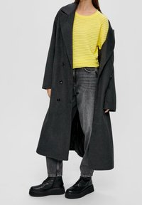 Lång grå dubbelknäppt kappa över en gul ribbad topp, i kombination med högmidjade mörka jeans och svarta plattformsskor.