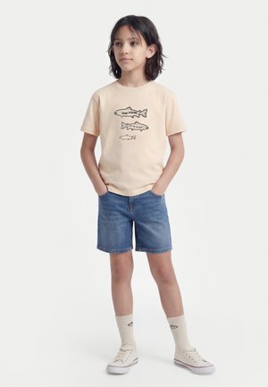 Enfant aux cheveux mi-longs portant un T-shirt beige avec des motifs de poisson, un short en denim, des chaussettes crème avec des poissons et des baskets crème.
