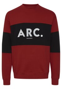 Rød og svart genser med rund hals, prydet med en iøynefallende hvit "ARC." logo og teksten "SOLID ARCHIVE" på forsiden. Blandet bomullsstoff.