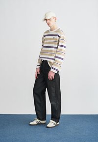 Camisola de manga comprida às riscas em bege e púrpura, combinada com jeans pretos de perna larga e ténis brancos com detalhes pretos. Boné creme usado.