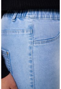 Jean en denim bleu clair avec une texture lisse, présentant des coutures surpiquées, des poches latérales et une ceinture élastique pour le confort.
