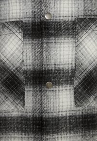 Sorte og hvide ternet skjorte med en tekstureret flannelstof. To store lommer med sølvfarvede metaltrykknapper på forsiden.