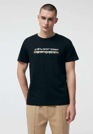 Młody mężczyzna o krótkich włosach, ubrany w czarną koszulkę z napisem "diverse sport" i beżowe spodnie, stoi na tle jednolitej białej ściany.