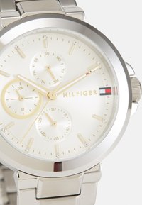Montre en acier inoxydable argenté avec un cadran crème, trois sous-cadrans, accents dorés et marqueurs en cristal. Présente un bracelet en métal poli.
