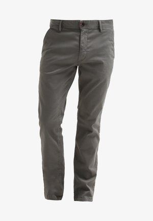 Mann, der ein blau kariertes Hemd mit Knöpfen, graue Hosen und schwarze hochgeschnittene Turnschuhe trägt und auf weißem Hintergrund geht.