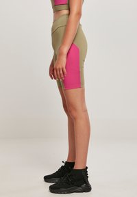 Biciklističke kratke hlače s visokim strukom u maslinasto zelenoj i ružičastoj boji, s glatkom teksturom i kontrastnim bočnim panelima. Nošene uz crne gležnjače.