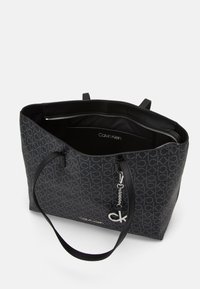Sac fourre-tout noir avec motif monogramme CK, texture lisse, compartiment zippé, intérieur noir et accents en hardware argenté.
