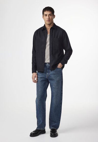 Manlig modell står självsäkert iklädd en svart skjorta över en grå linne och blå löst sittande jeans, med svarta skor.