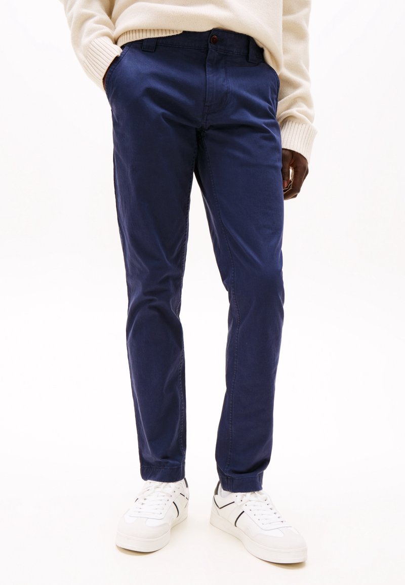 Homme portant un chino bleu marine, des baskets blanches et un pull crème, debout avec une main dans la poche sur fond blanc.
