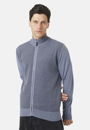 Uomo con capelli scuri che indossa un maglione grigio strutturato con zip e pantaloni neri, in piedi su uno sfondo semplice.