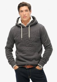 Sudadera gris con un diseño de logo texturizado, cordones crema, bolsillo delantero, puños acanalados, combinada con jeans oscuros y un reloj.
