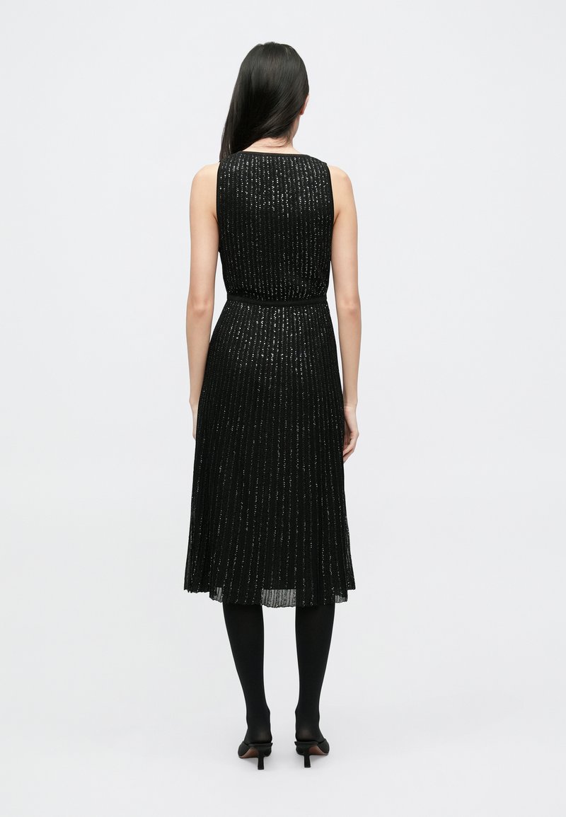 Black pleated sleeveless dress with shiny thread details, cinched waist, and knee-length skirt, paired with black tights and heels.