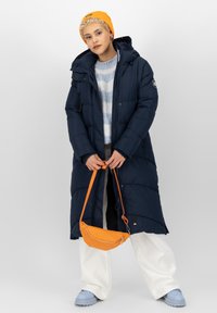 Giacca a vento blu navy con cappuccio, con chiusura a zip e tasche. Abbinata a un maglione a strisce blu chiaro, pantaloni bianchi e una borsa arancione.