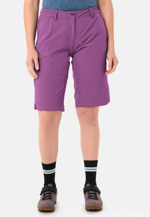 Vrouw draagt paarse knielange shorts, zwarte sokken met witte strepen en taupekleurige sportschoenen, staat tegen een effen achtergrond.