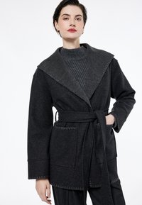 Abrigo de mezcla de lana en gris oscuro con un gran cuello, cintura ceñida con cinturón y detalles con costuras. Cuenta con dos bolsillos delanteros y una capa inferior tejida texturizada.