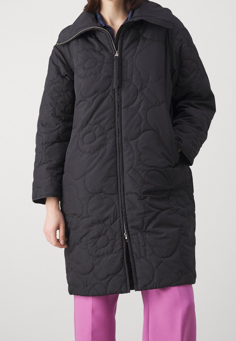 Black quilted outerwear with floral pattern, high collar, and zipper closure, paired with light pink trousers. Smooth texture and lightweight design.