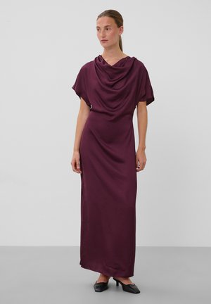 Femme debout portant une longue robe drapée violet profond à manches courtes et des chaussures à talons noires sur un fond gris clair uni.