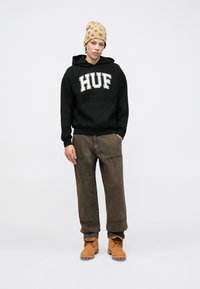 Black hoodie with "HUF" printed in white, oversized fit, paired with loose brown pants and tan boots. A beige polka dot beanie adds texture.