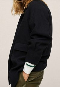 Laivaston sininen villasekoitetakki väljällä istuvuudella, teksturoidulla kankaalla ja sivutaskuilla. Hihansuissa valkoiset ja vihreät raidat.