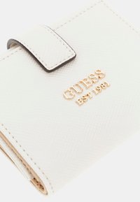 Baltas tekstūruotas piniginė su auksiniu "GUESS" logotipu ir relifiniu užrašu "EST 1981", turinti kontrastingą tamsią dirželio užsegimą.