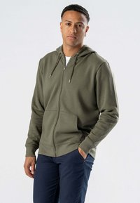 Olivengrøn zip-up hættetrøje lavet af blødt stof med en frontlomme, justerbare snore og ribbede manchetter. Båret over en hvid t-shirt.