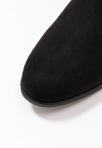 Zapato de ante negro con punta redondeada y textura suave. La suela es de goma negra, lo que proporciona un acabado flexible y duradero.