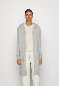 Hellgrauer langer Cardigan mit Kapuze, gerippten Bündchen und Fronttaschen. Stricktextur, offenes Design, getragen über einem beigen Oberteil und weißen Hosen.