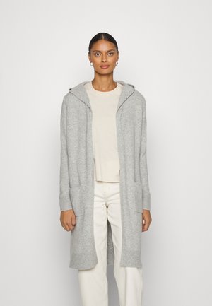 Cardigan long gris clair avec capuche, poignets côtelés et poches avant. Texture tricotée, design ouvert, porté sur un haut beige et un pantalon blanc.