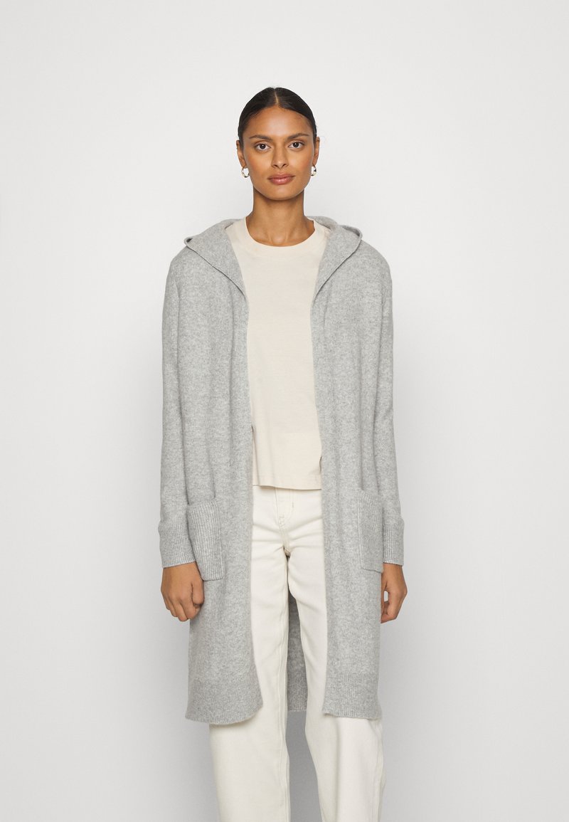 Hellgrauer langer Cardigan mit Kapuze, gerippten Bündchen und Fronttaschen. Stricktextur, offenes Design, getragen über einem beigen Oberteil und weißen Hosen.