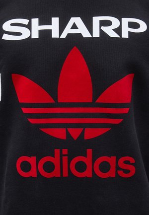 Musta kangas, jossa suuri punainen Adidas trefoil -logo ja teksti alapuolella, päällä valkoinen isoilla kirjaimilla kirjoitettu sana "SHARP" logon yläpuolella.